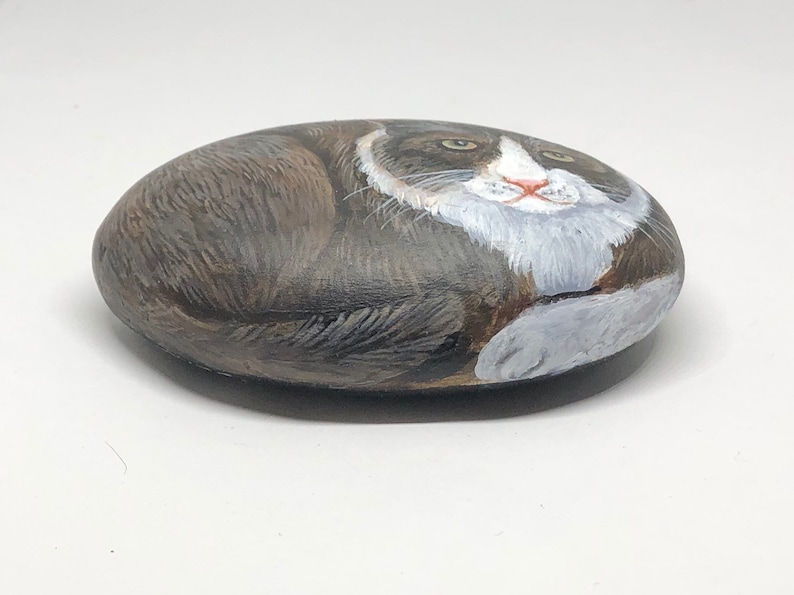 May include: Hand-painted rock art depicting a gray and white cat with green eyes. The cat is painted in a realistic style and is lying down on its side.
