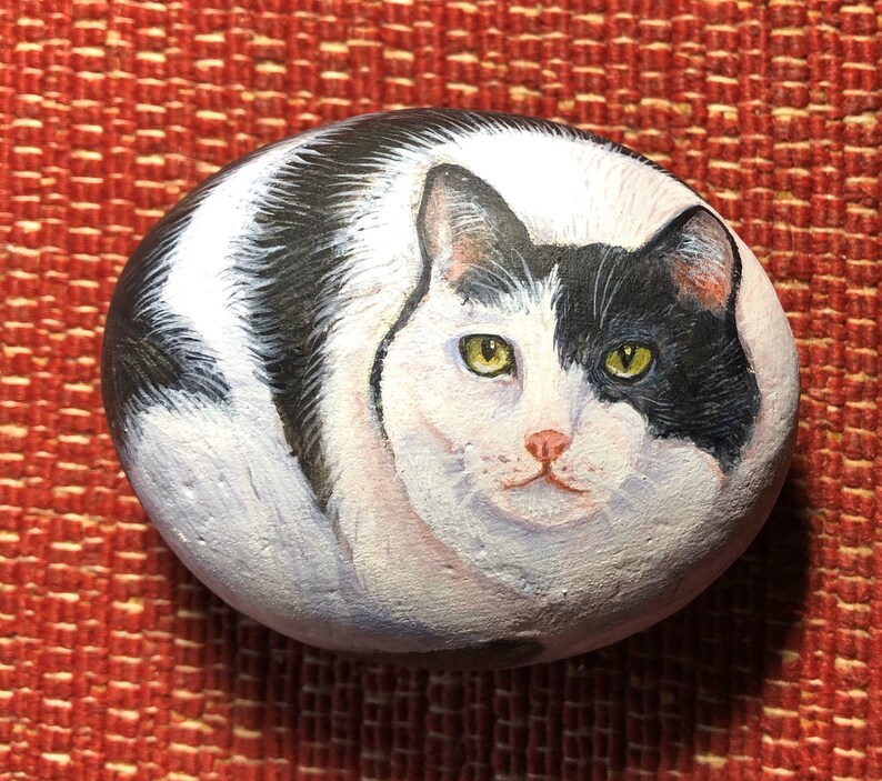 May include: Hand-painted rock art depicting a black and white cat with yellow eyes. The cat is curled up in a sleeping position. The rock is smooth and has a natural, rounded shape.