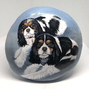 May include: Hand-painted rock art featuring two Cavalier King Charles Spaniels. The dogs are painted in realistic detail with black, white and brown fur. The background is a light blue colour.