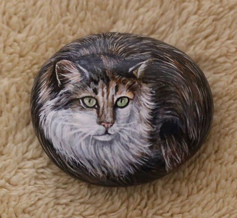 May include: Hand-painted rock art depicting a realistic portrait of a long-haired cat with green eyes. The cat is painted in shades of brown, white, and black.
