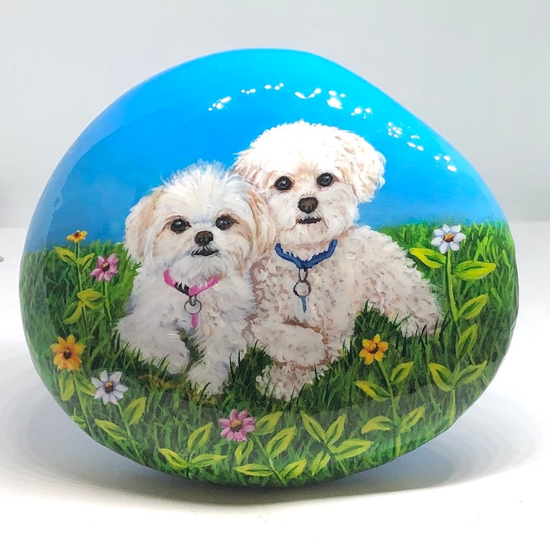May include: Hand-painted rock art featuring two white dogs with blue and pink collars, sitting in a field of green grass and colourful flowers against a blue sky.