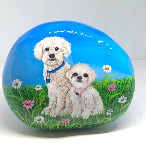 May include: Hand-painted rock featuring two white dogs with blue and pink collars, sitting in a field of green grass and colourful flowers.