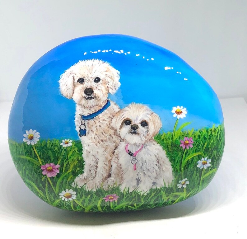 May include: Hand-painted rock featuring two white dogs with blue and pink collars, sitting in a field of green grass and colourful flowers.