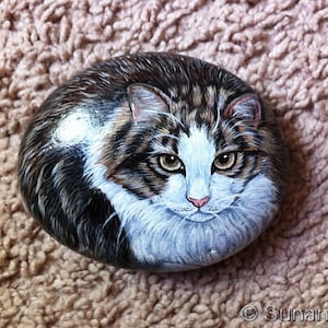 May include: A hand-painted rock depicting a tabby cat with white markings. The cat has green eyes and a pink nose. The rock is painted realistically and has a smooth, rounded surface.