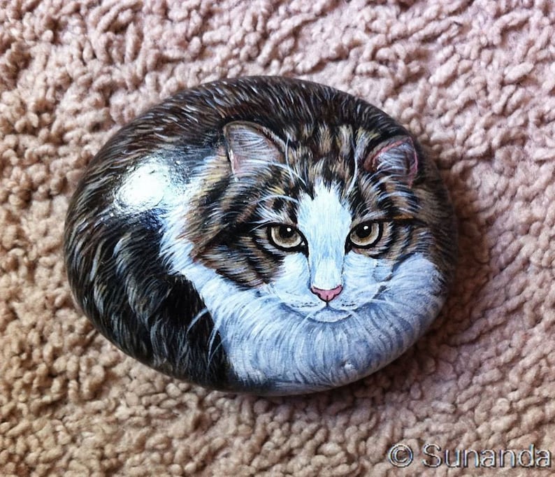 May include: A hand-painted rock depicting a tabby cat with white markings. The cat has green eyes and a pink nose. The rock is painted realistically and has a smooth, rounded surface.