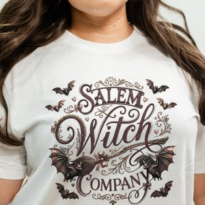 May include: White t-shirt featuring a Salem Witch Company design. The design includes bats, a broom, and the words "Salem Witch Company" in a decorative font. The design is in shades of brown and purple.