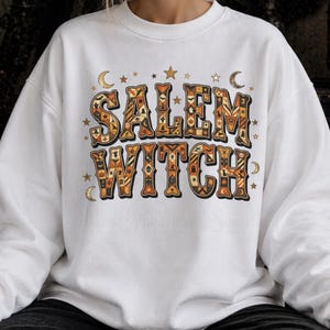 Puede incluir: Una sudadera blanca de cuello redondo con el texto SALEM WITCH en letras grandes y negritas. Las letras están rellenas con un patrón repetitivo de iconos de Halloween en naranja, negro y crema, incluyendo murciélagos, calabazas y estrellas.