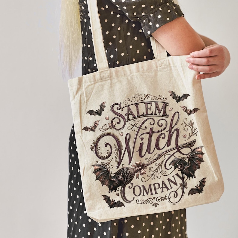May include: A cream-colored tote bag featuring the words "Salem Witch Company" in a decorative font, with bats and a broom graphic. The bag is held by a person wearing a black and white polka dot dress.