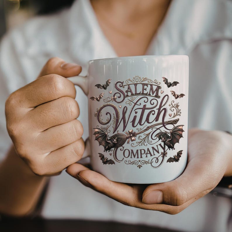 May include: White ceramic mug featuring the words "Salem Witch Company" in a decorative script, with bat and broom illustrations. The mug is held in both hands.