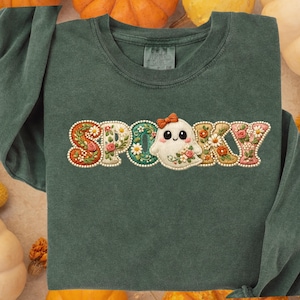 Può includere: Una maglietta a maniche lunghe verde scuro con la scritta "SPOOKY" ricamata sul davanti. Le lettere sono decorate con motivi floreali e un personaggio fantasma. La maglietta è circondata da zucche.