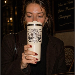 May include: A cream-colored travel mug with the text "Salem Witch Company" in a gothic font, with bat illustrations. The mug is held by a person with red nail polish and gold rings. The background is blurred, suggesting a cafe setting.