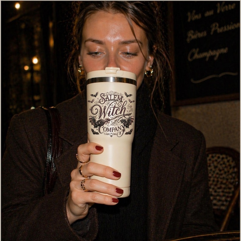May include: A cream-colored travel mug with the text "Salem Witch Company" in a gothic font, with bat illustrations. The mug is held by a person with red nail polish and gold rings. The background is blurred, suggesting a cafe setting.
