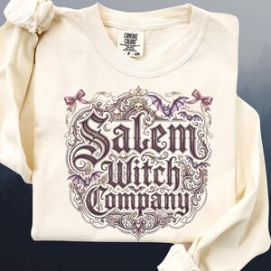 May include: Cream-colored long-sleeve shirt featuring the text "Salem Witch Company" in a decorative, gothic-style font. The design includes bats, skulls, and floral elements in shades of purple and pink. The shirt is made of a soft material.