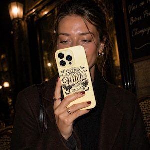 May include: A pale yellow phone case with the words "Salem Witch Company" in a gothic font, along with bat and witch-themed graphics. The case has a camera cutout and is held in front of a person.