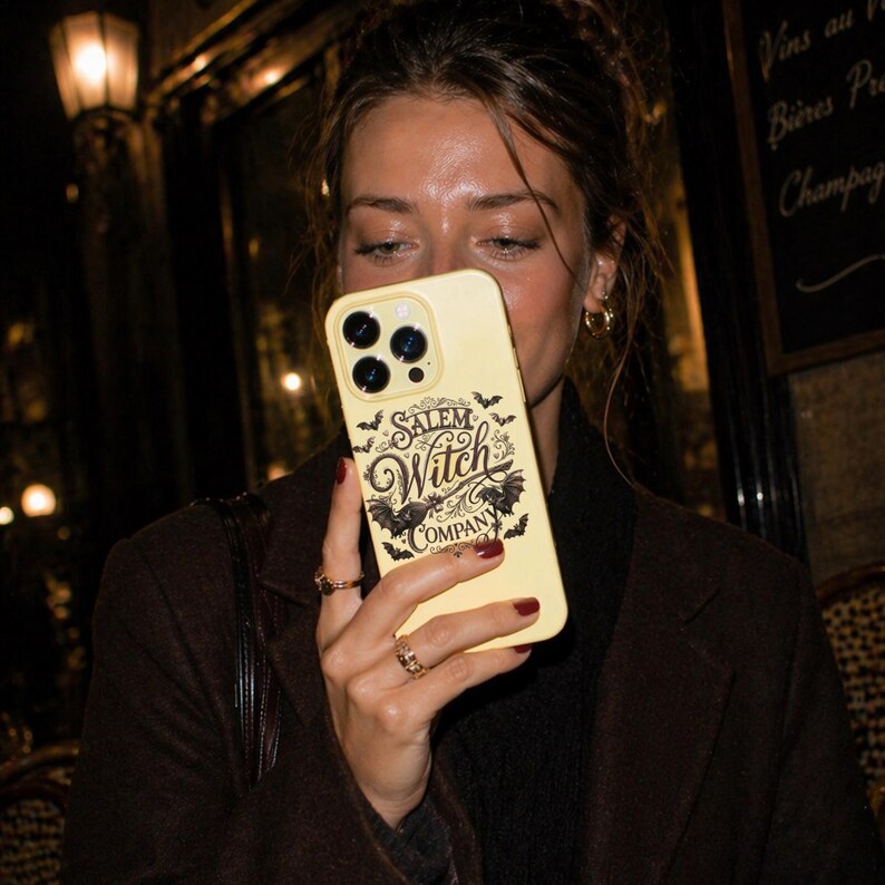 May include: A pale yellow phone case with the words "Salem Witch Company" in a gothic font, along with bat and witch-themed graphics. The case has a camera cutout and is held in front of a person.