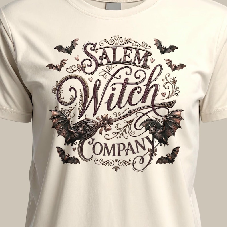 May include: Cream-colored t-shirt featuring the words "Salem Witch Company" in a decorative, vintage-style font. The design includes bats and a broom, with a gothic aesthetic. The shirt is a crew neck.