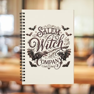 May include: Cream-colored spiral-bound notebook featuring the text "Salem Witch Company" in an ornate, vintage-style font. The design includes bats and a witch's broom. The notebook has a black spiral binding.