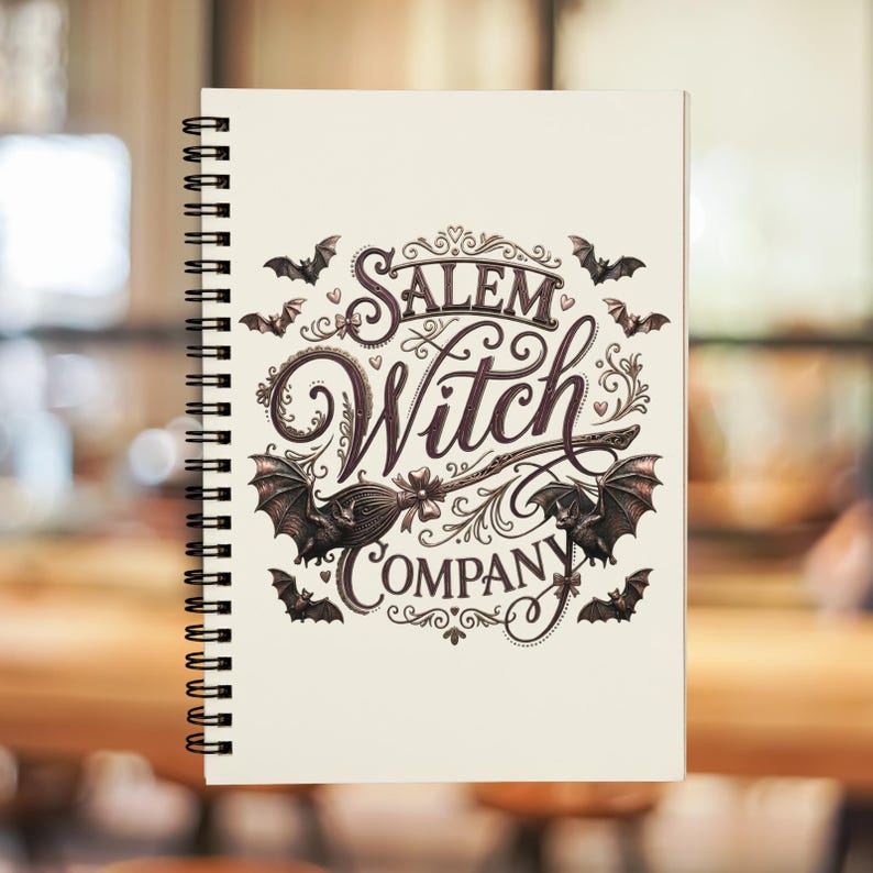 May include: Cream-colored spiral-bound notebook featuring the text "Salem Witch Company" in an ornate, vintage-style font. The design includes bats and a witch's broom. The notebook has a black spiral binding.
