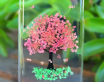 May include: A clear phone case featuring a pressed flower design of a tree. The tree is pink and green, with a black silhouette, and surrounded by pink and green flowers.