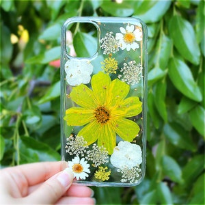 May include: A clear phone case with a yellow flower and white flowers pressed inside. The case has a gold glitter effect.
