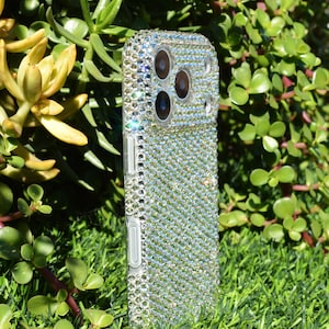 Puede incluir: Una funda transparente para teléfono incrustada con brillantes pedrería iridiscente. La funda tiene recortes para la cámara y cubiertas para los botones. La funda del teléfono está rodeada de hierba verde y follaje.