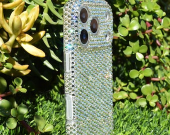 May include: A clear phone case encrusted with sparkling, iridescent rhinestones. The case has a camera cutout and button covers. The phone case is surrounded by green grass and foliage.