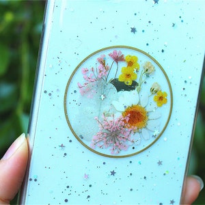May include: A light blue phone case with a clear circle containing pressed flowers and gold glitter. The background of the case is covered in silver glitter and stars.
