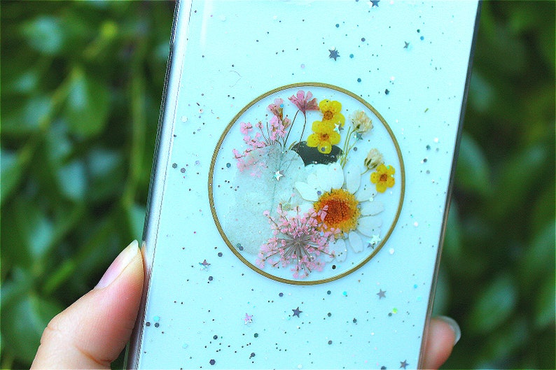May include: A light blue phone case with a clear circle containing pressed flowers and gold glitter. The background of the case is covered in silver glitter and stars.