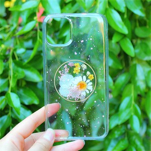 May include: A clear phone case with a glittery background and a pressed flower design in a circle. The flowers are white, yellow, and pink.