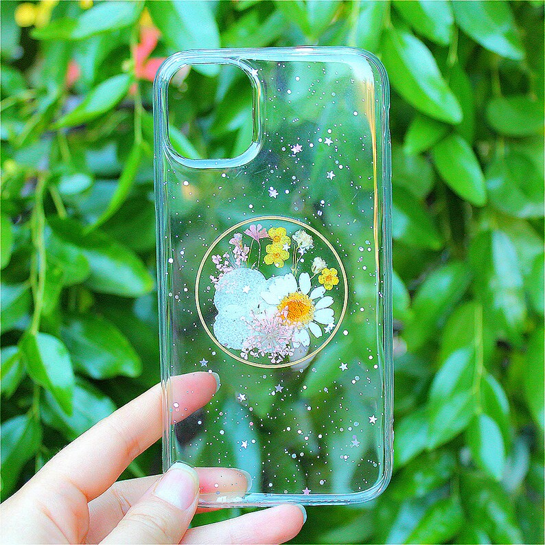 May include: A clear phone case with a glittery background and a pressed flower design in a circle. The flowers are white, yellow, and pink.