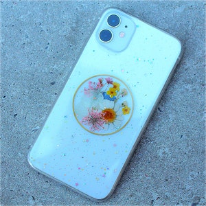 May include: A white phone case with a clear, glittery background and a round, gold-rimmed, pressed flower design.