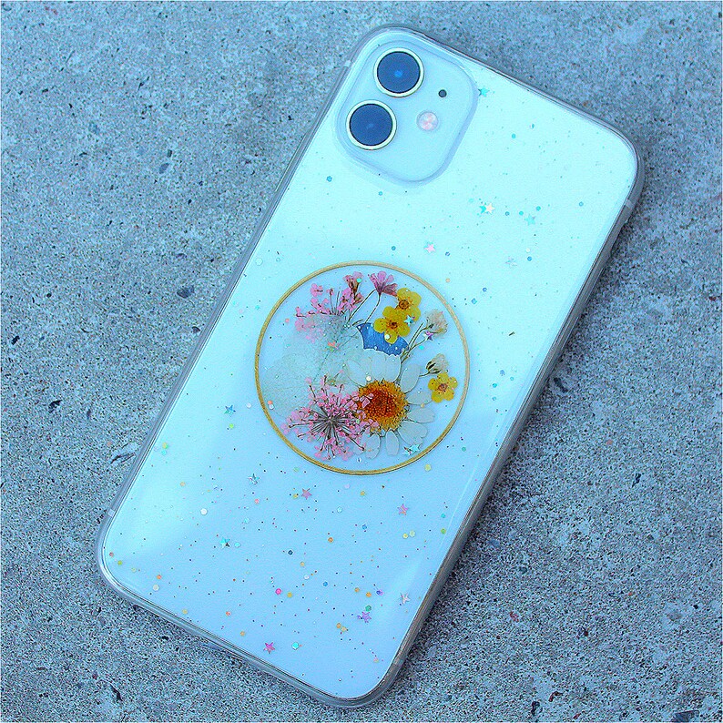 May include: A white phone case with a clear, glittery background and a round, gold-rimmed, pressed flower design.