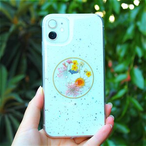 May include: A light blue phone case with a clear, round sticker featuring pressed flowers. The sticker has a white background and features a variety of yellow, white, and pink flowers.
