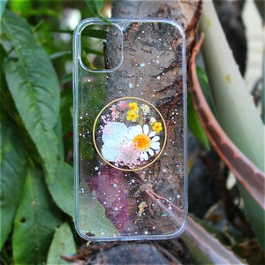 May include: A clear phone case with a glittery background and a pressed flower design in a circle. The flowers are white, yellow, and pink.