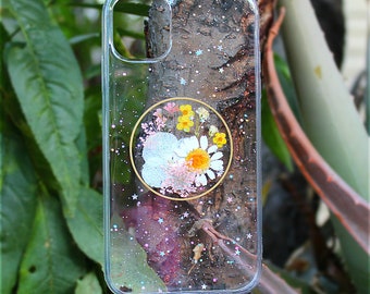 May include: A clear phone case with a glittery background and a pressed flower design in a circle. The flowers are white, yellow, and pink.