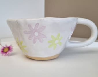 May include: A white ceramic mug with a light pink and yellow floral design. The mug has a handle and a slightly rounded shape.