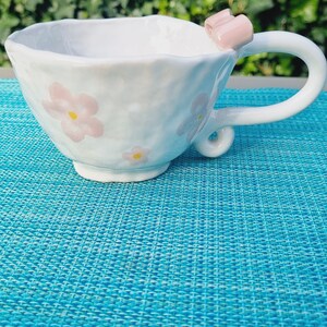 May include: White ceramic teacup with a pink bow handle and floral design. The cup is sitting on a blue textured surface.