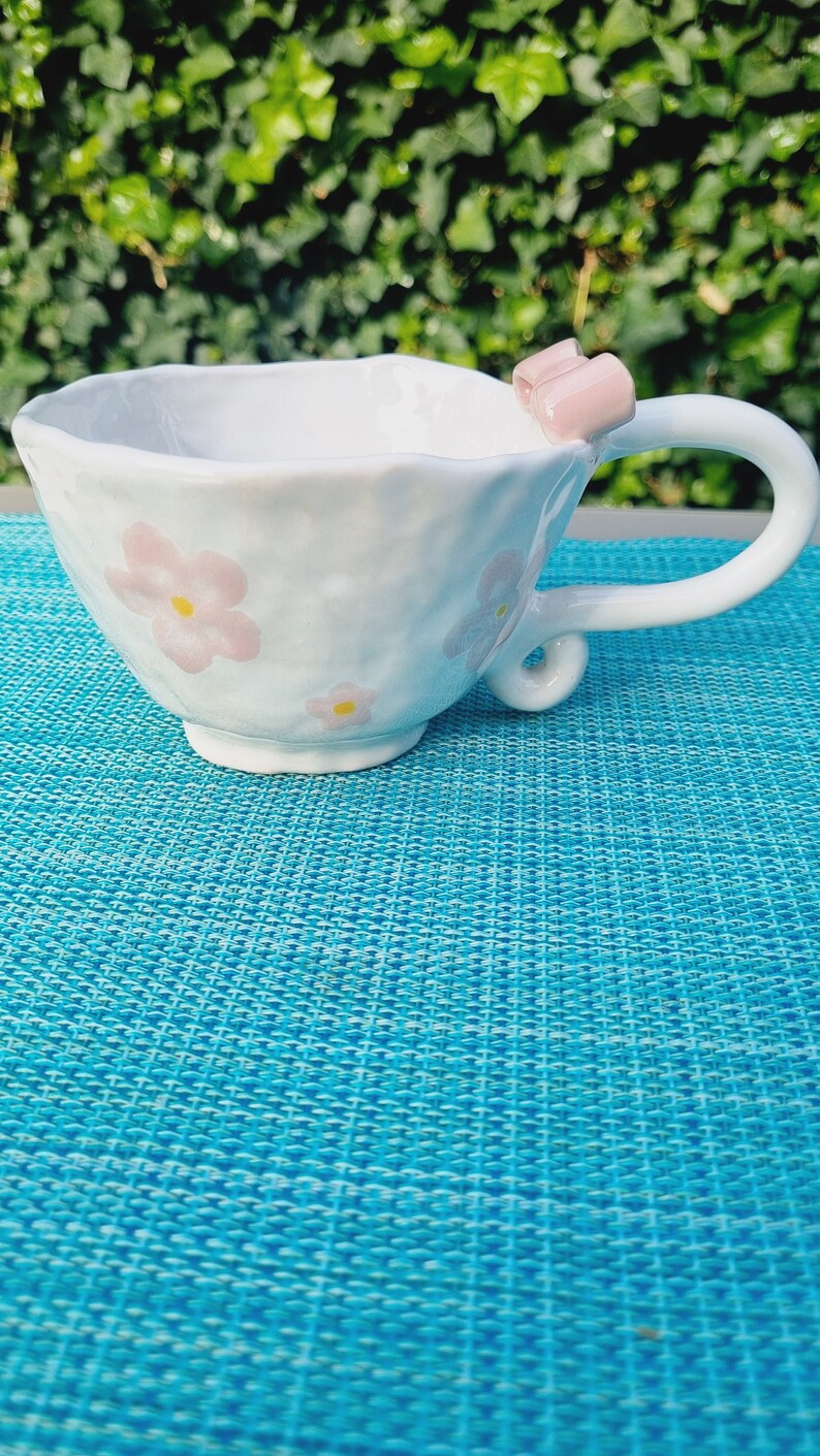 May include: White ceramic teacup with a pink bow handle and floral design. The cup is sitting on a blue textured surface.