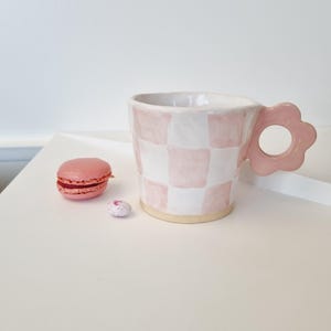 May include: A ceramic mug with a pink and white checkered pattern. The mug has a flower-shaped handle and a light pink base. A pink macaron and a small candy are next to the mug. The mug is on a white surface.