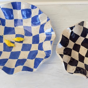 May include: Two ceramic plates with a wavy edge. One plate is blue and white checkered, and the other is black and white checkered. The plates have a speckled, off-white background. Yellow dried flowers are placed on the blue and white plate.