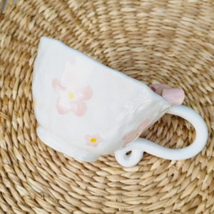 May include: White ceramic teacup with a pink floral design. The cup has a unique handle that is shaped like a flower.