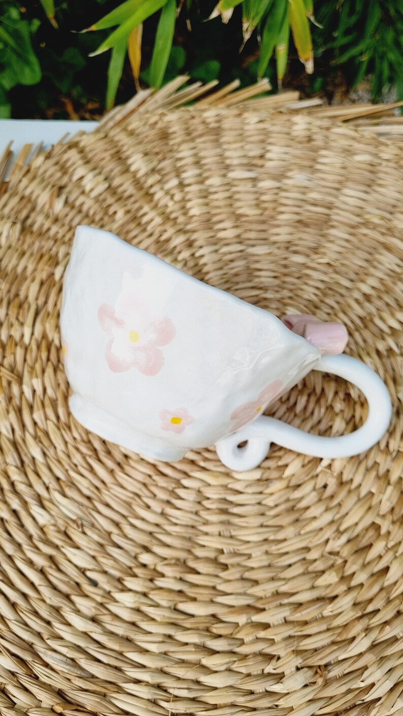 May include: White ceramic teacup with a pink floral design. The cup has a unique handle that is shaped like a flower.