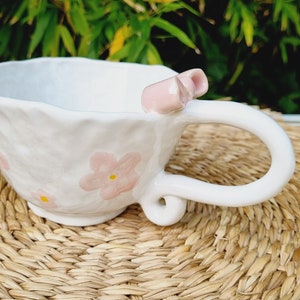 May include: White ceramic teacup with a pink bow and pink floral designs. The cup is sitting on a woven straw mat.
