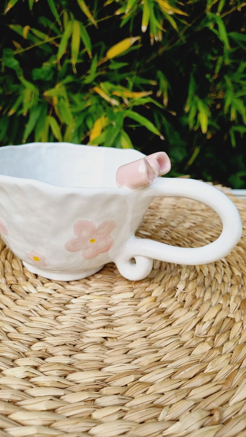 May include: White ceramic teacup with a pink bow and pink floral designs. The cup is sitting on a woven straw mat.