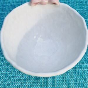 May include: White ceramic bowl with a pink bow on the top. The bowl is sitting on a blue and white patterned surface.