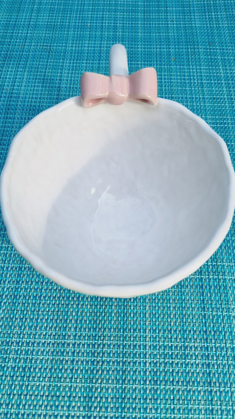 May include: White ceramic bowl with a pink bow on the top. The bowl is sitting on a blue and white patterned surface.