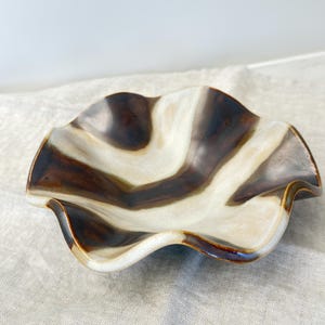 May include: A ceramic bowl with a wavy, organic shape. The bowl features a brown and cream-colored pattern, with the colors swirling across the surface. The rim has a dark brown edge. The bowl is likely used for serving or display.