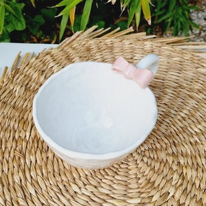 May include: White ceramic bowl with a pink bow handle. The bowl is sitting on a woven straw mat.
