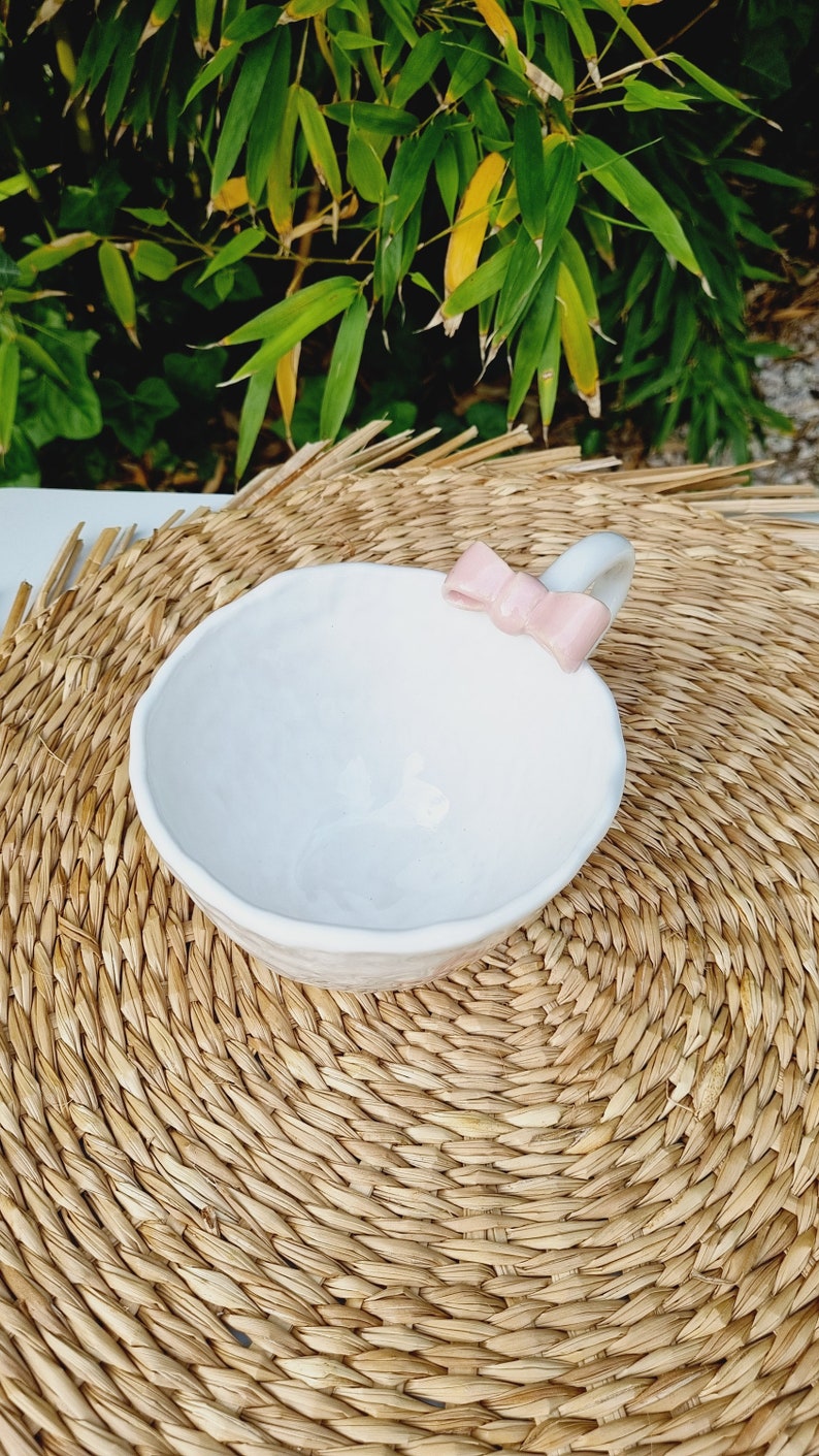 May include: White ceramic bowl with a pink bow handle. The bowl is sitting on a woven straw mat.