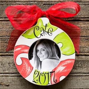 May include: A round, white ceramic ornament with a photo of a person. The ornament is decorated with red and green swirls and the name "Cate" and the year "2017". A red ribbon is tied at the top.